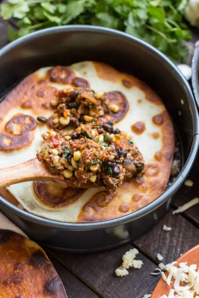 A wood spoon adding taco meat with beans and corn to a tortilla in a springform pan.