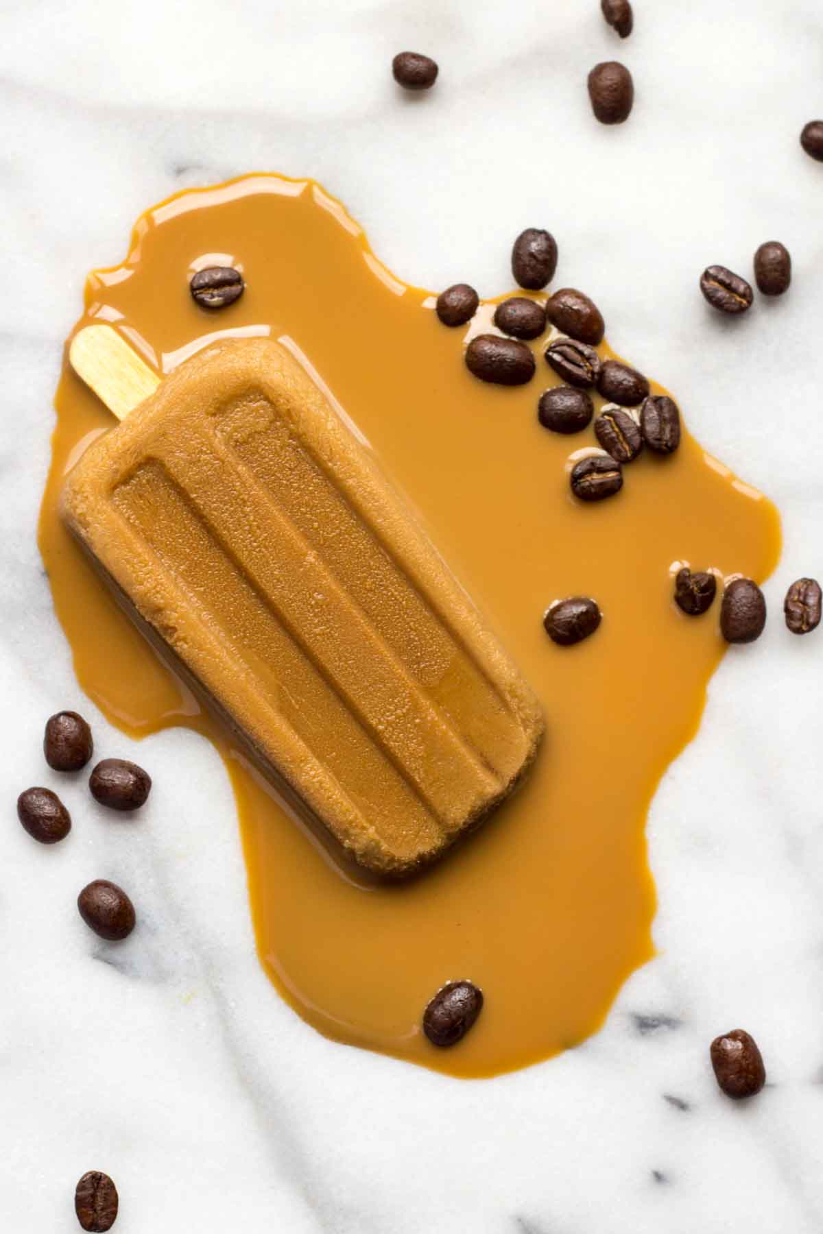 Melted coffee popsicle on a white marble background surrounded by scattered roasted coffee beans, with coffee pooling dramatically around the base-perfect for a summer dessert concept.