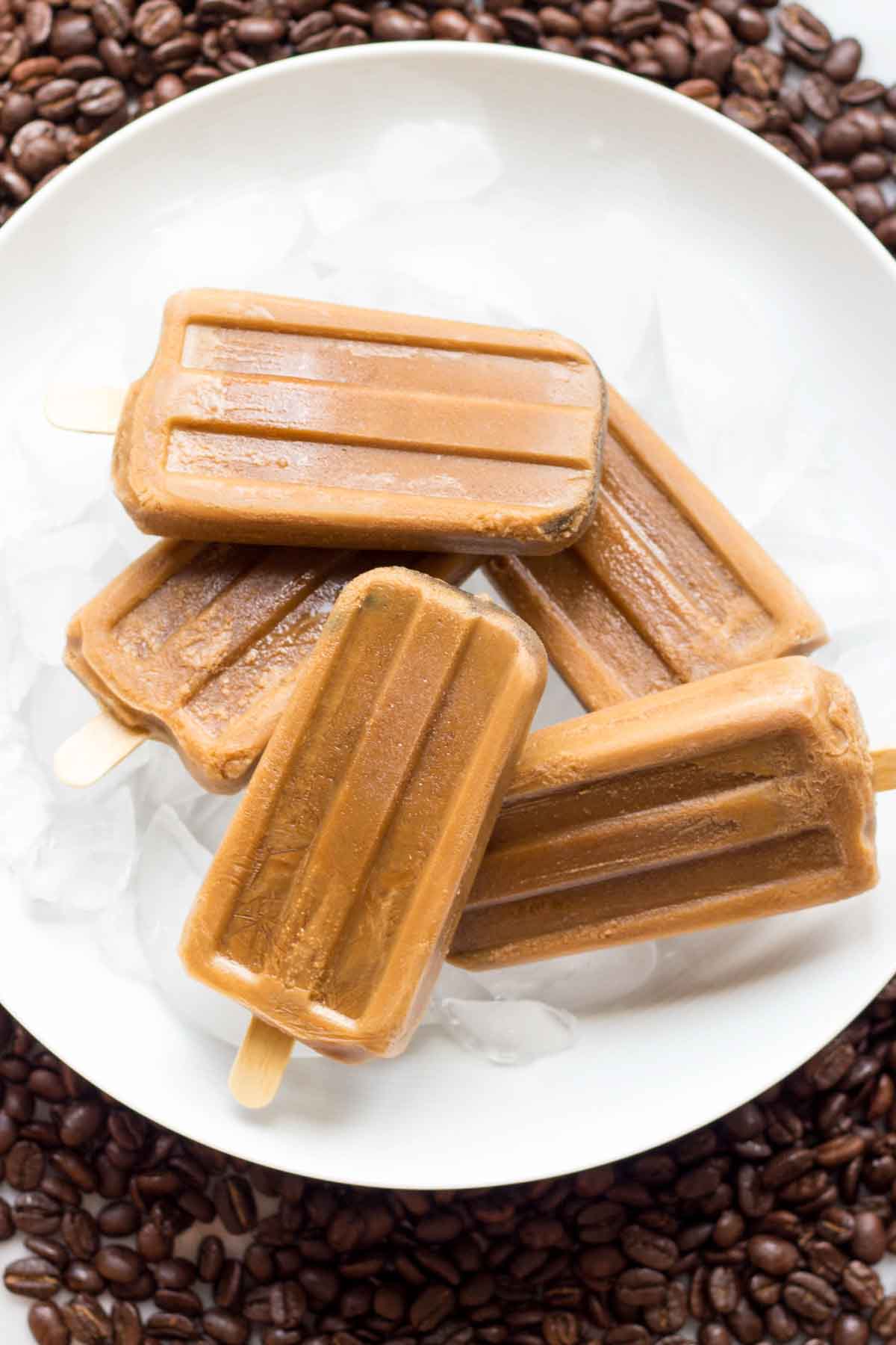 Coffee popsicles stacked in a white bowl filled with ice, placed on a ring of coffee beans-showing a refreshing and caffeinated frozen treat ideal for hot days.