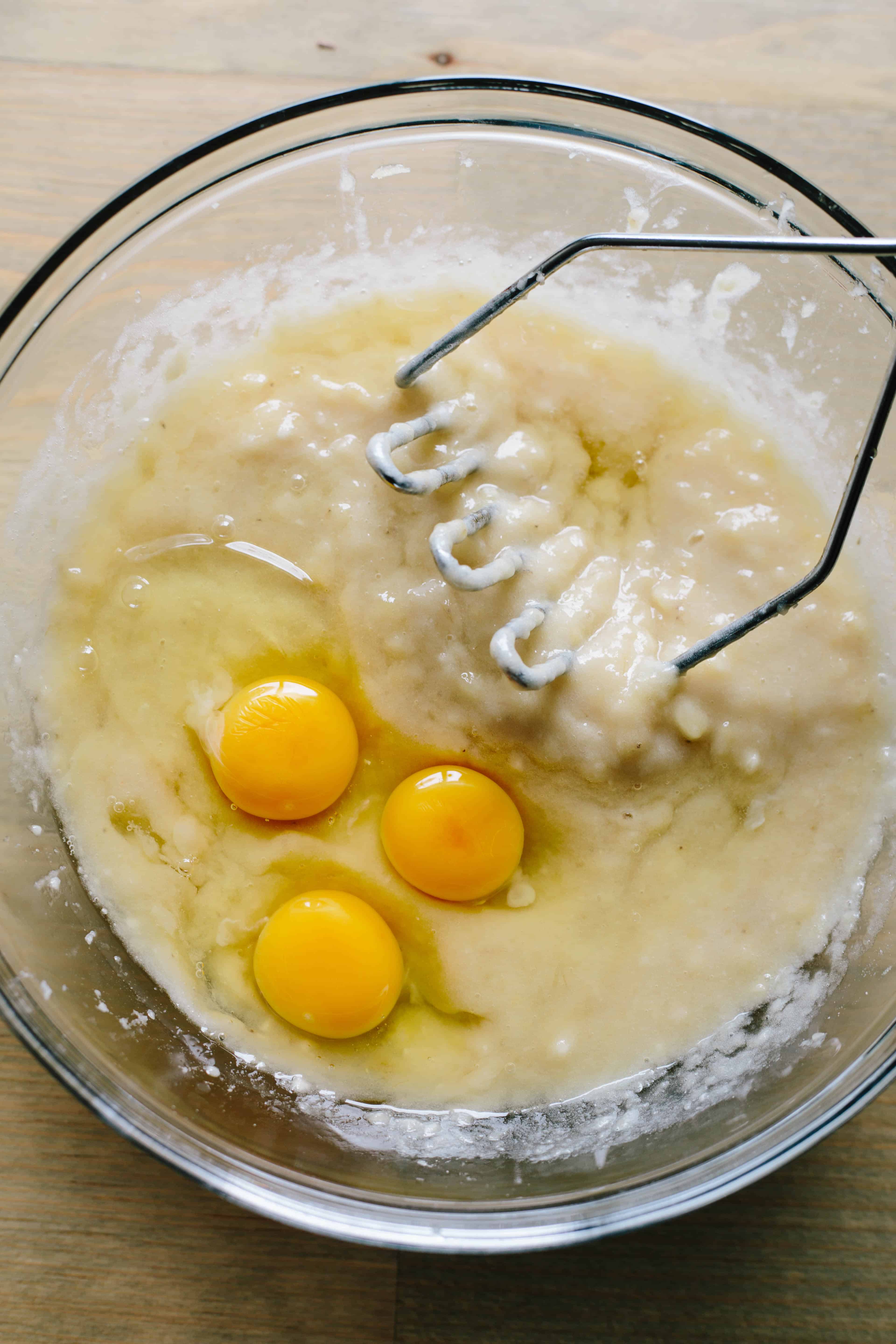 A mixing bowl of banana bread batter including three eggs and a potato masher.