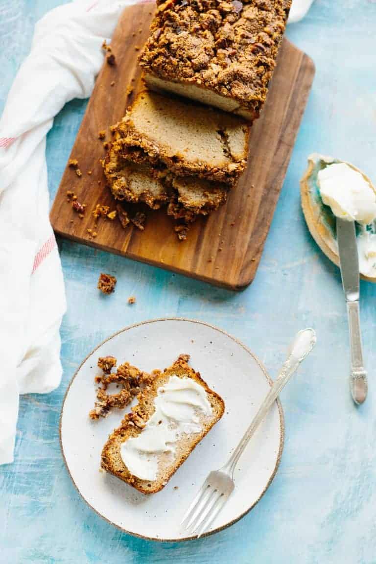 Loaf of Paleo banana bread sliced on a wood cutting board, next to a plate containing a buttered slice.