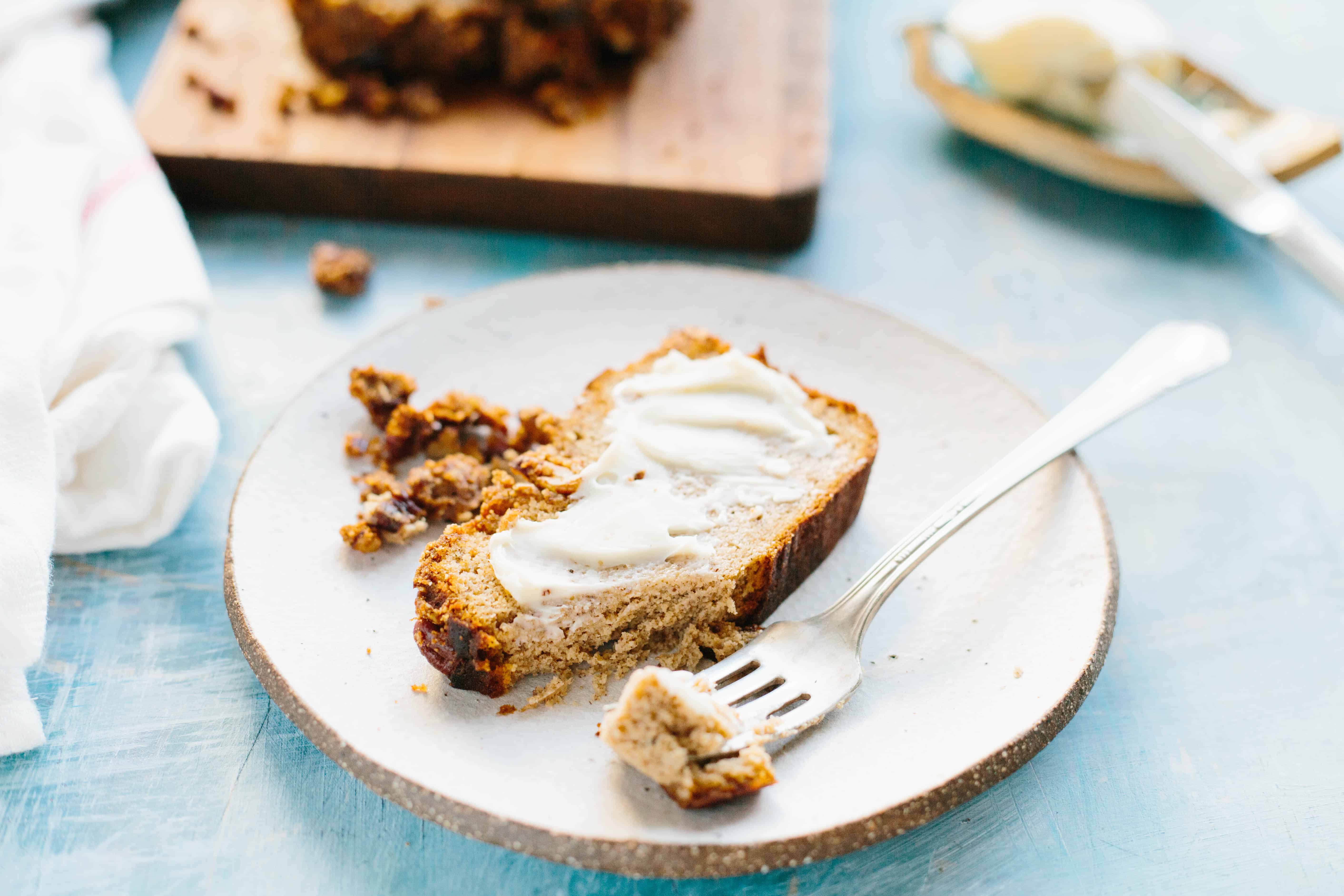 A fork with a bite of banana bread on it resting on a plate next to the rest of the slice.