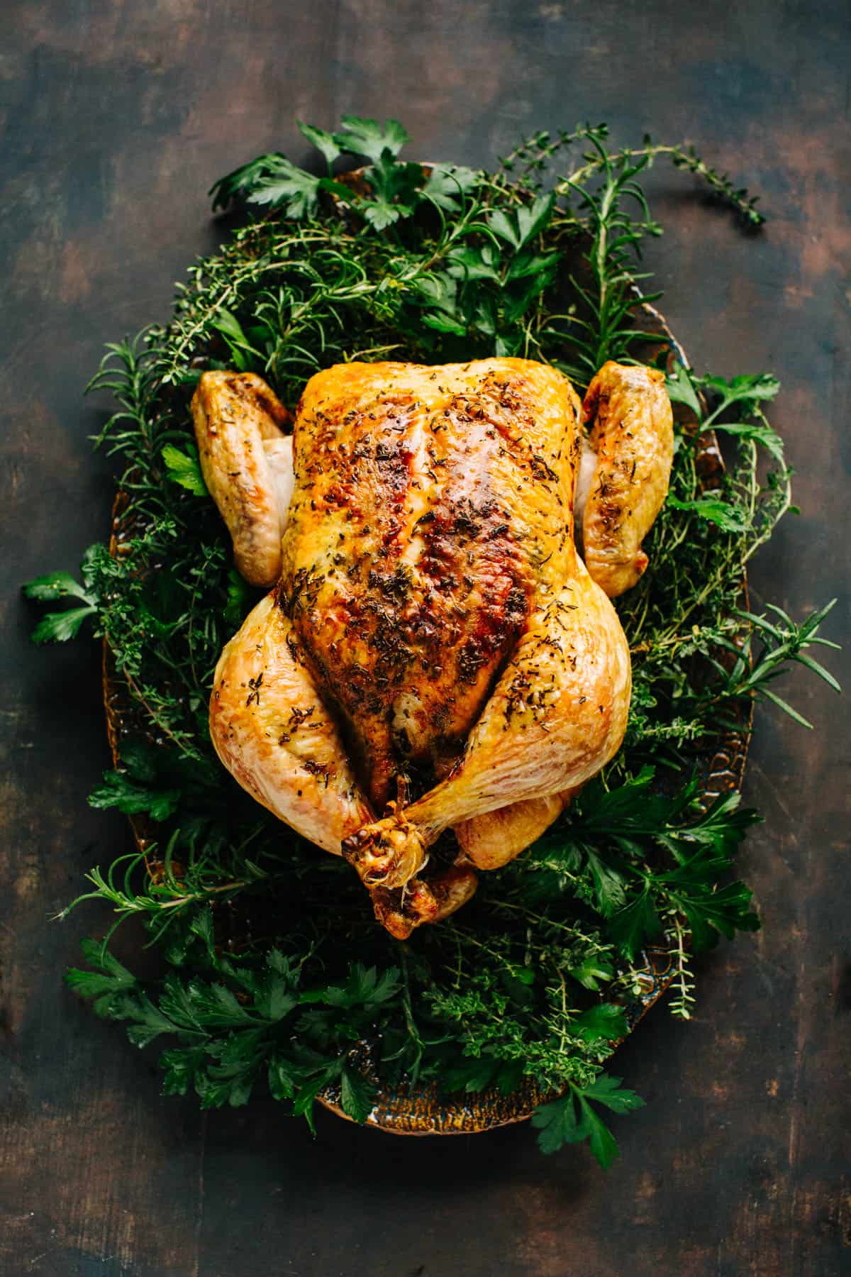 whole roasted chicken on a serving platter surrounded by fresh herbs on dark wood background