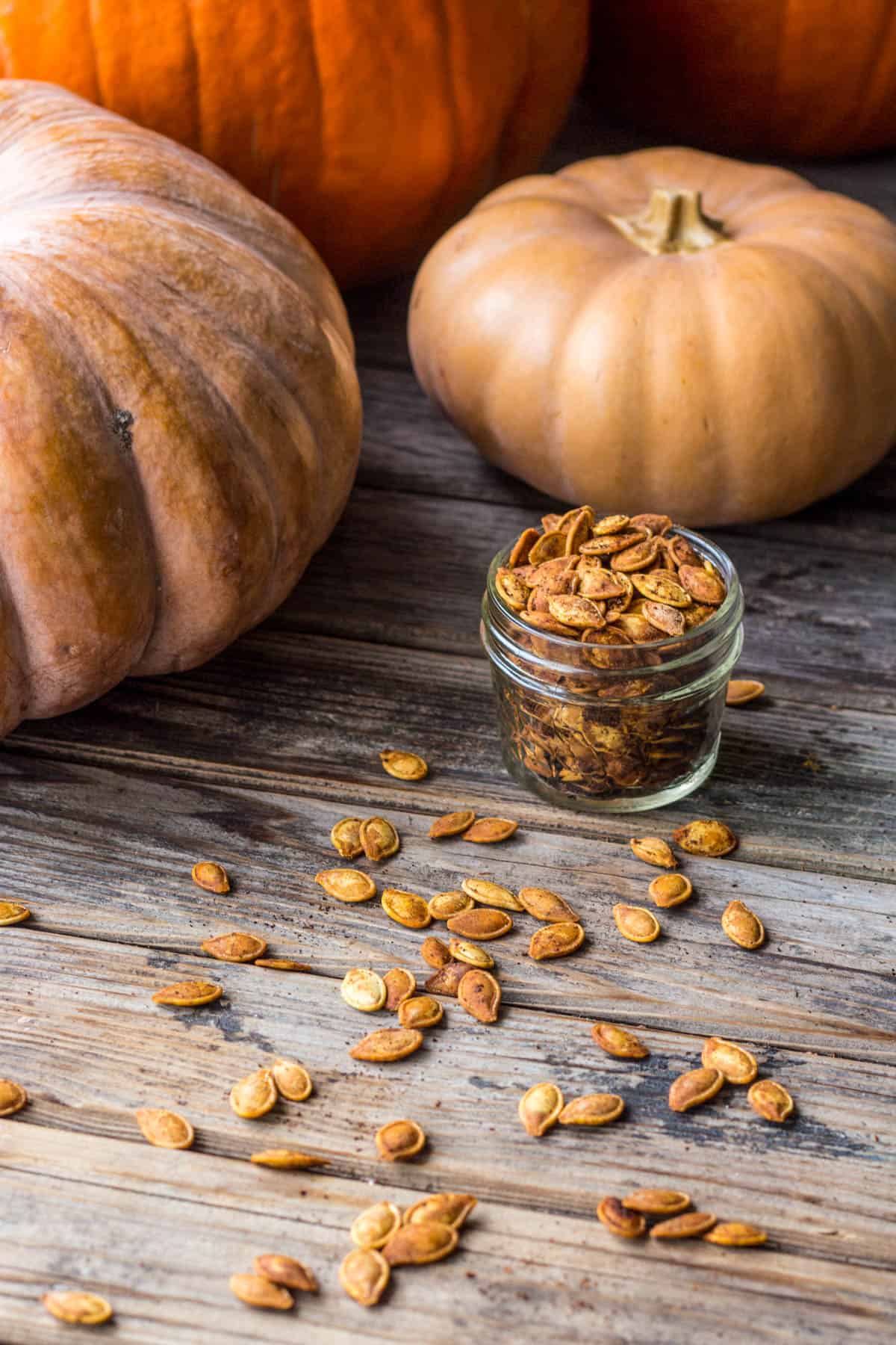 small jar of roasted pumpkin seeds on wood table with seeds scattered around and tan cinderella pumpkins in the background