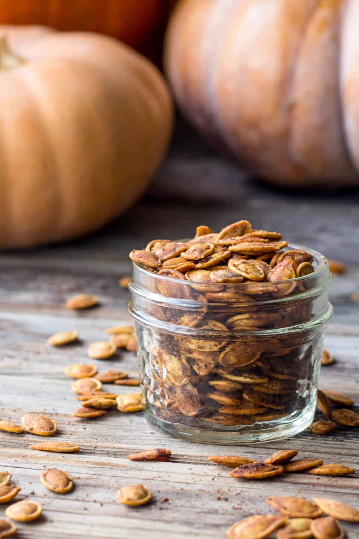 close up sideview of small jar of roasted pumpkin seeds on wood table with seeds scattered in the foreground and assorted pumpkins in the background