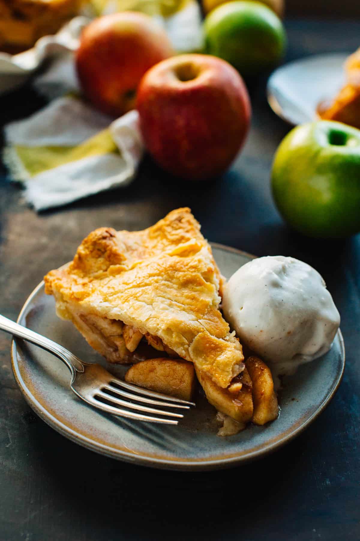 slice of apple pie on a gray plate with a fork and scoop of ice cream with apples in the background
