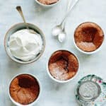Three mini chocolate souffles in ramekins next to a bowl of freshly whipped cream.