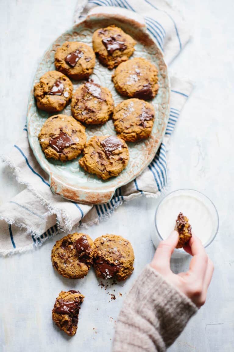 A hand dunking a chocolate chip cookie into a glass of milk next to a serving platter of more cookies.