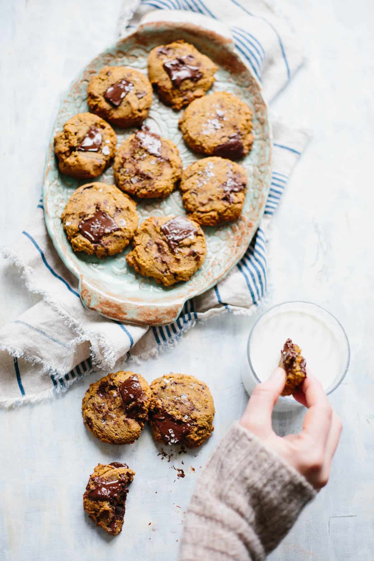 A hand dunking a Paleo chocolate chip cookie into a glass of milk next to a serving platter of more cookies.