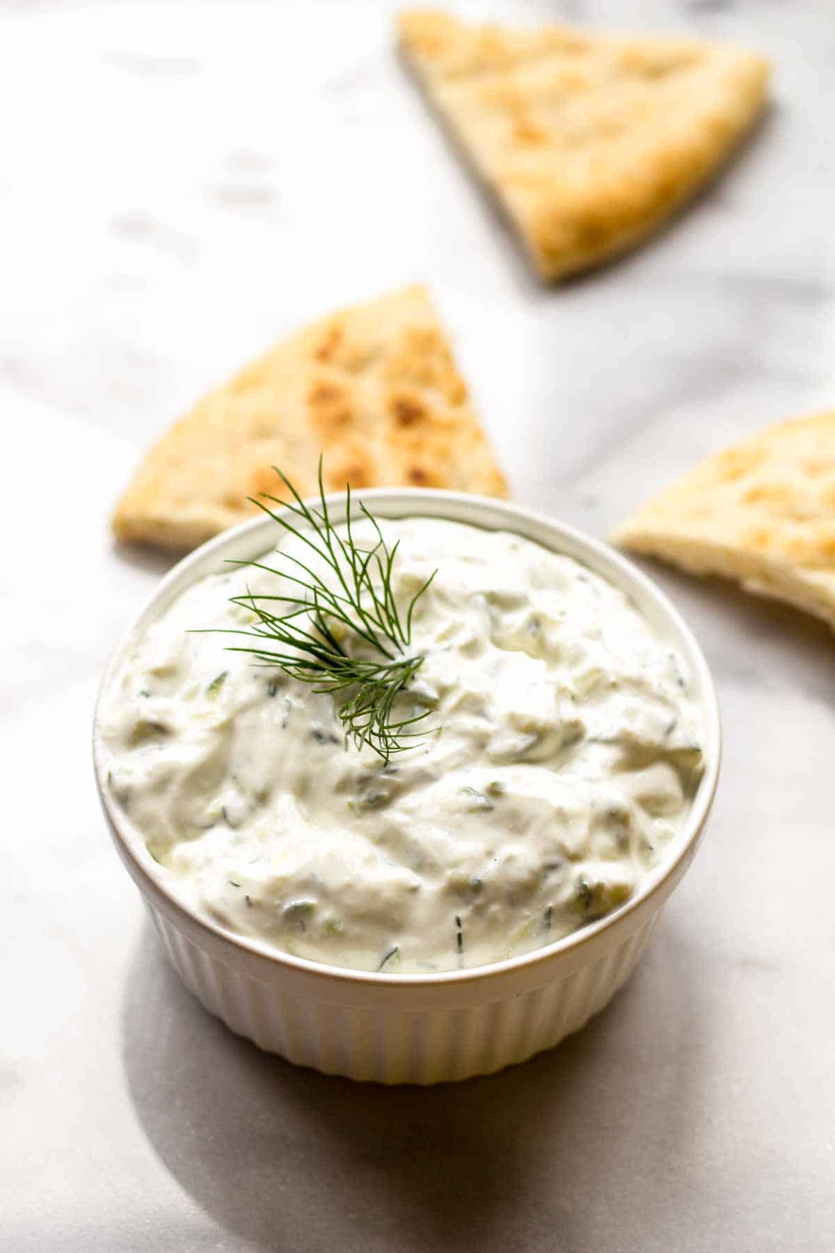 A white ramekin of Tzatziki Sauce with pita triangles in the background.