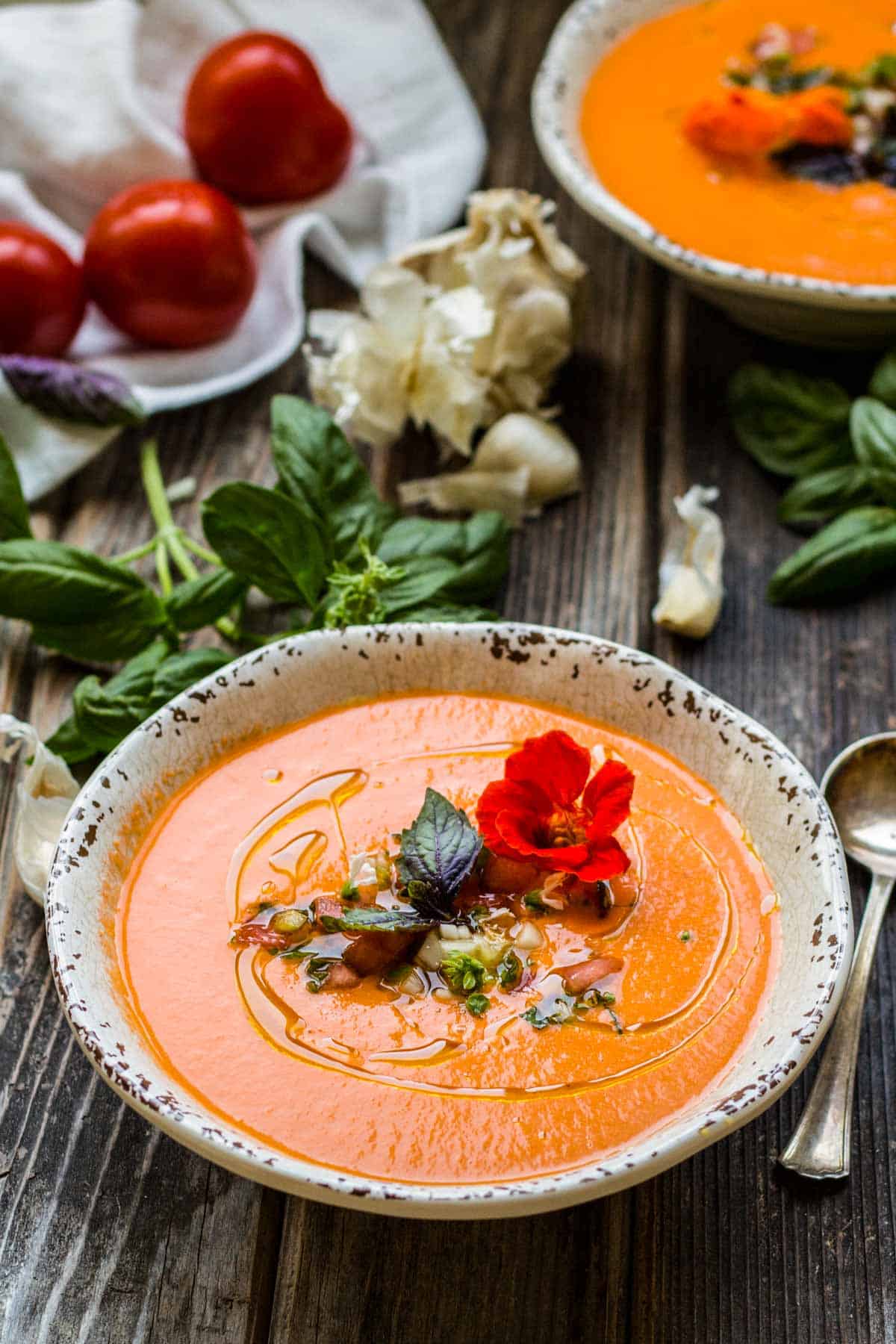 A wooden table with two bowls of gazpacho along with some raw ingredients.