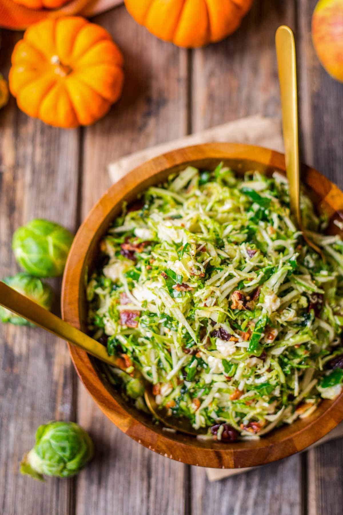 Overhead shot of a big wooden bowl of salad with pumpkins and whole Brussels sprouts in the background.