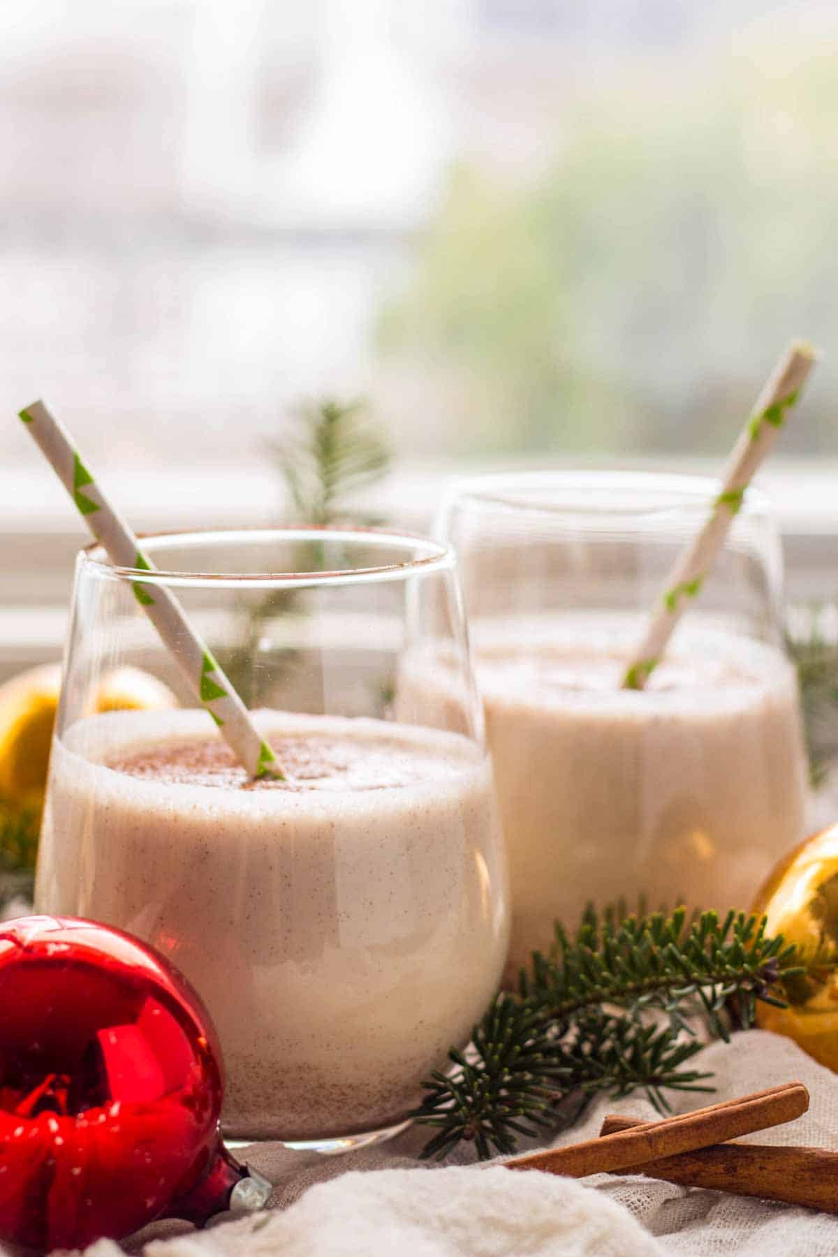 Side view of two glasses of Coquito with straws in front of a window.