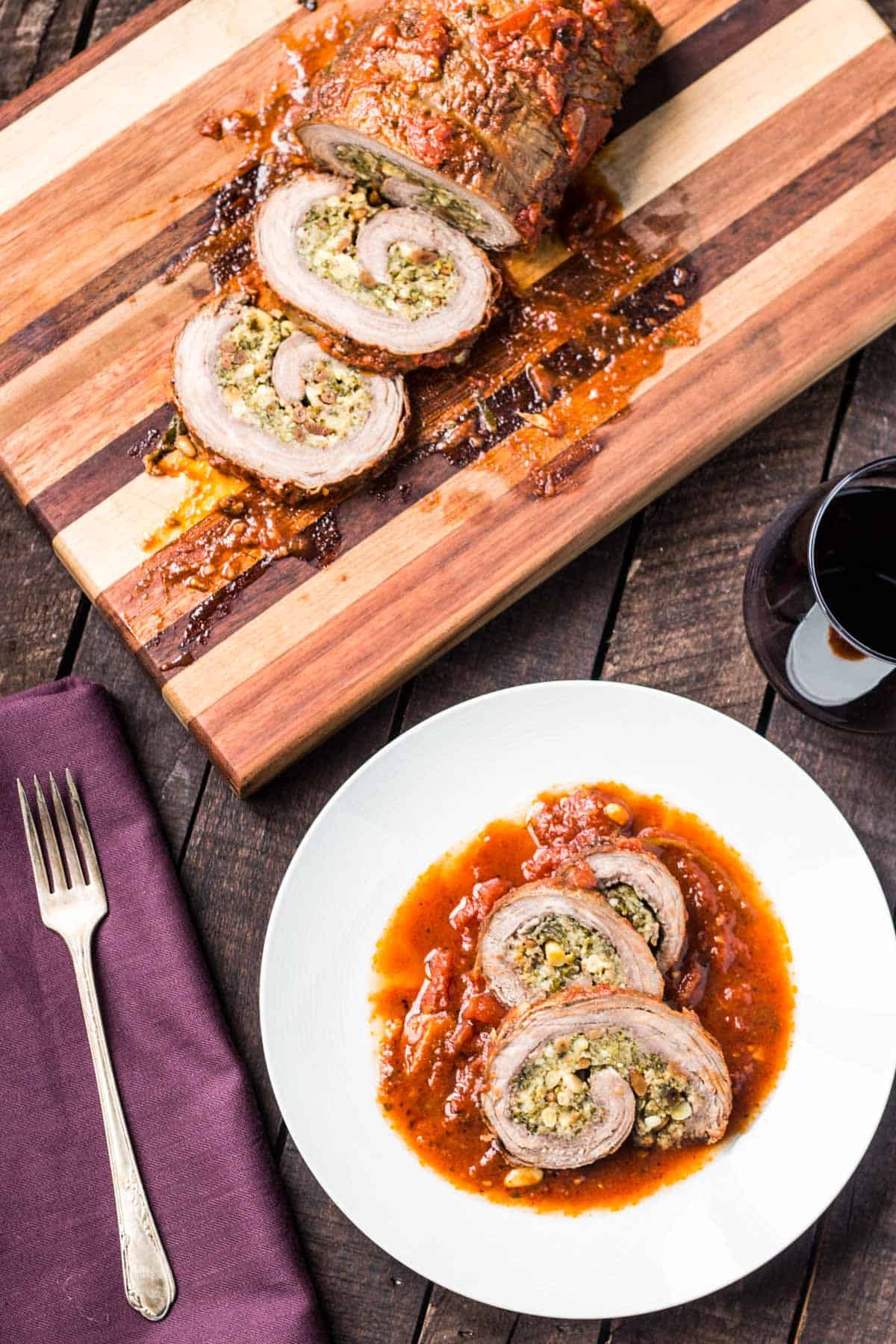 A dark wooden table with a cutting board and plate with braciole on them along with a purple napkin, fork and a glass of red wine.