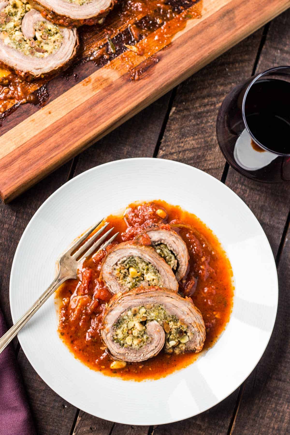 Overhead shot of a plate of beef braciole on a dark wood table with a glass of red wine.