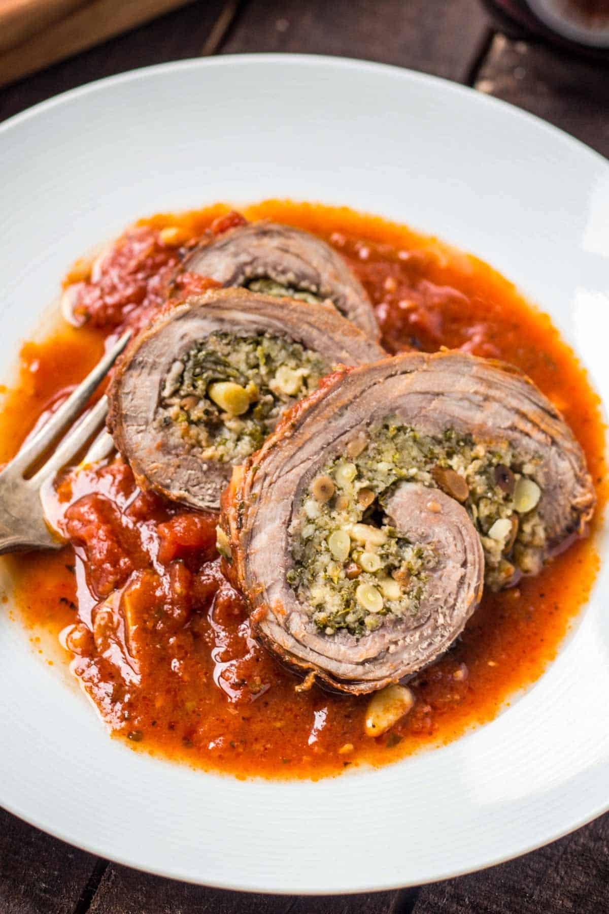 Close up of 3 slices of braciole over red sauce on a white plate with a fork.
