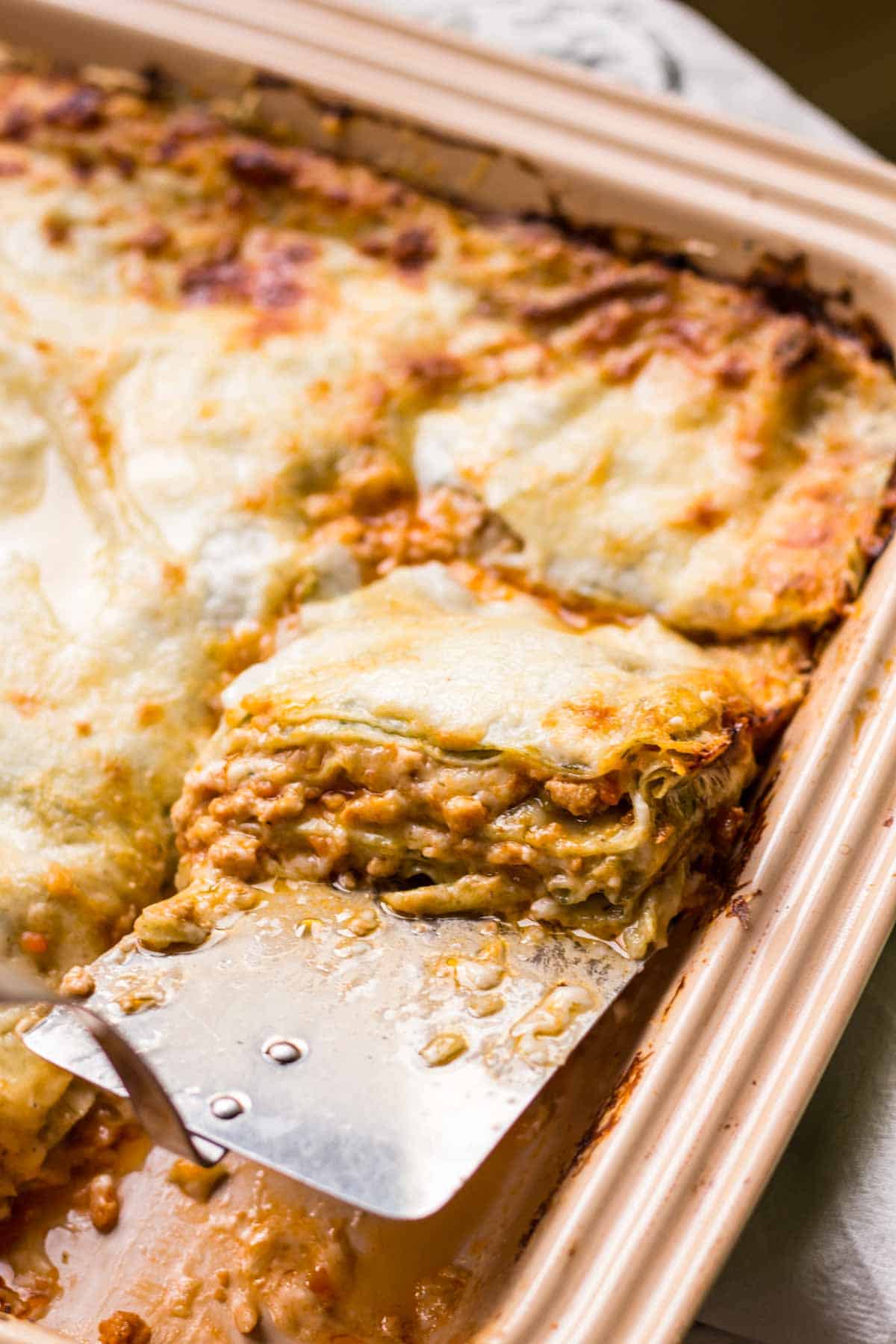 A spatula lifting a slice of lasagna bolognese from a stoneware baking dish.