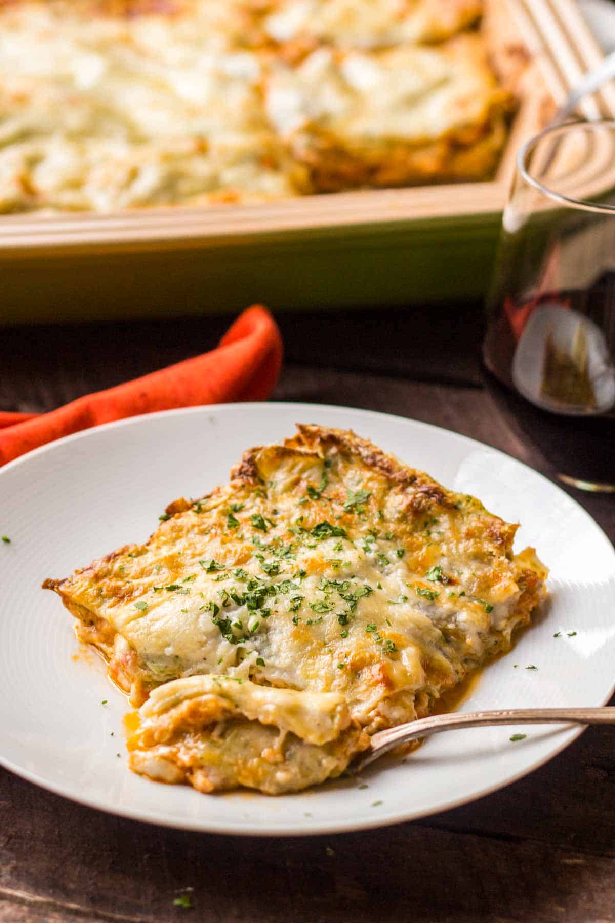 Vertical shot of a slice of lasagna bolognese on a white plate with a fork with the baking dish in the background.