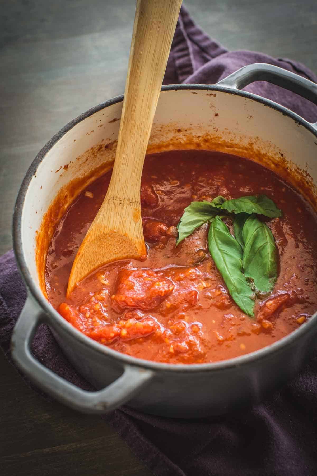 A pot of homemade marinara sauce with a wooden spoon and sprig of basil.