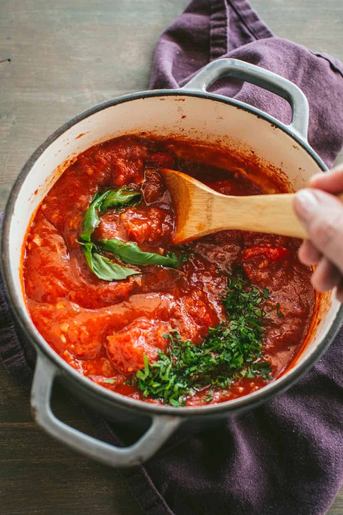 A hand holding a wooden spoon stirring herbs into a pot of tomato sauce.