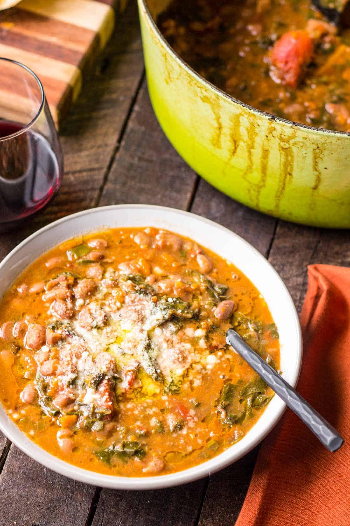 A bowl of bean soup with a spoon, a glass of red wine and a green Dutch oven in the background.