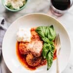 Overhead shot of a white plate with meatballs, a scoop of ricotta, fresh basil and a glass of red wine.