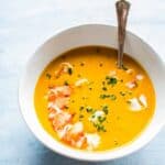 A white bowl with bright orange lobster soup with a lobster tail and chives on top.