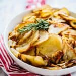 A white bowl of roasted potatoes with a sprig of rosemary on top and a red napkin.