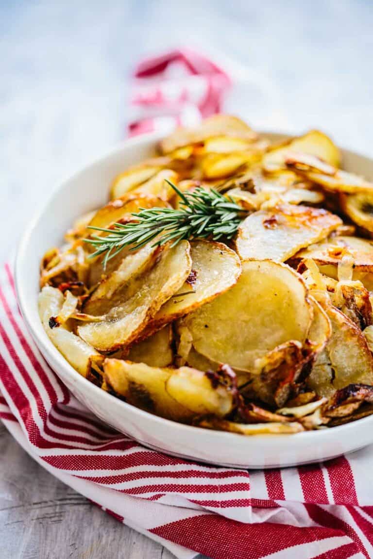 A white bowl of roasted potatoes with a sprig of rosemary on top and a red napkin.