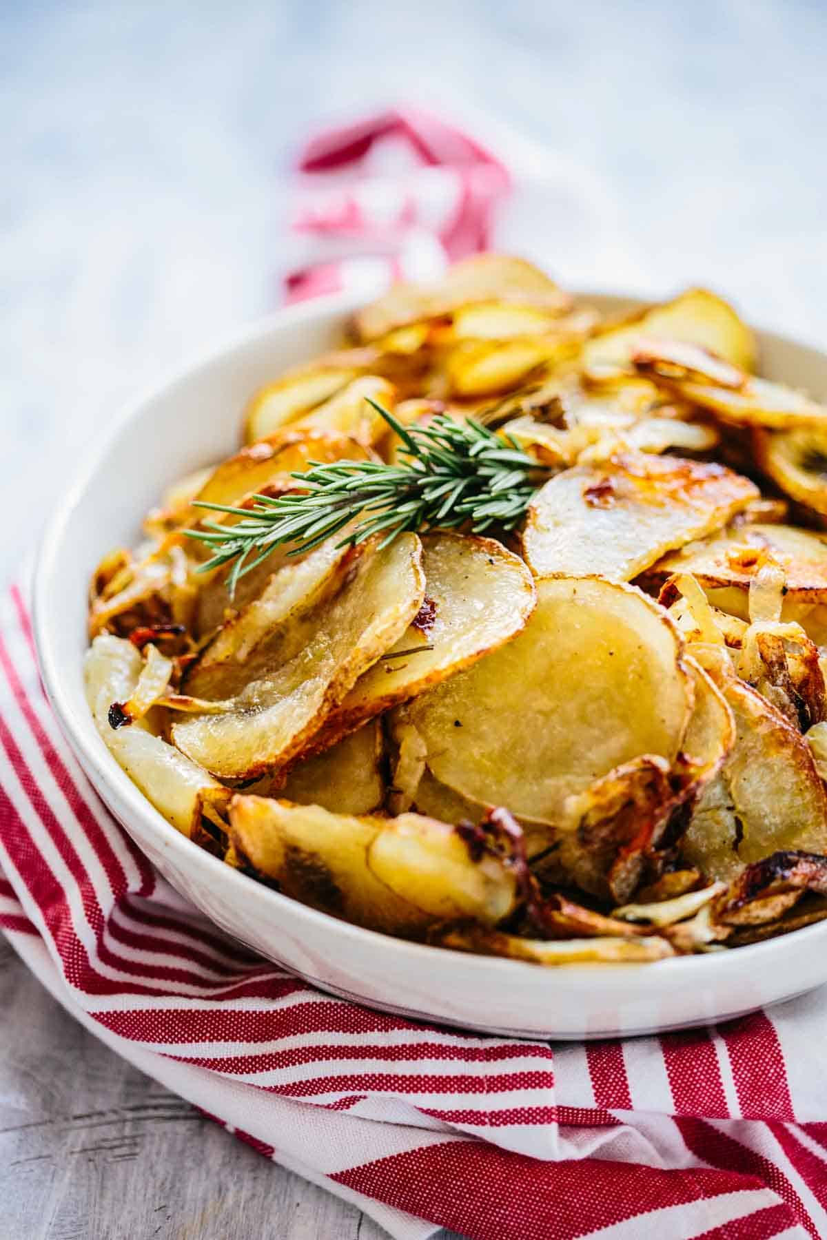 A white bowl of roasted potatoes with a sprig of rosemary on top and a red napkin.