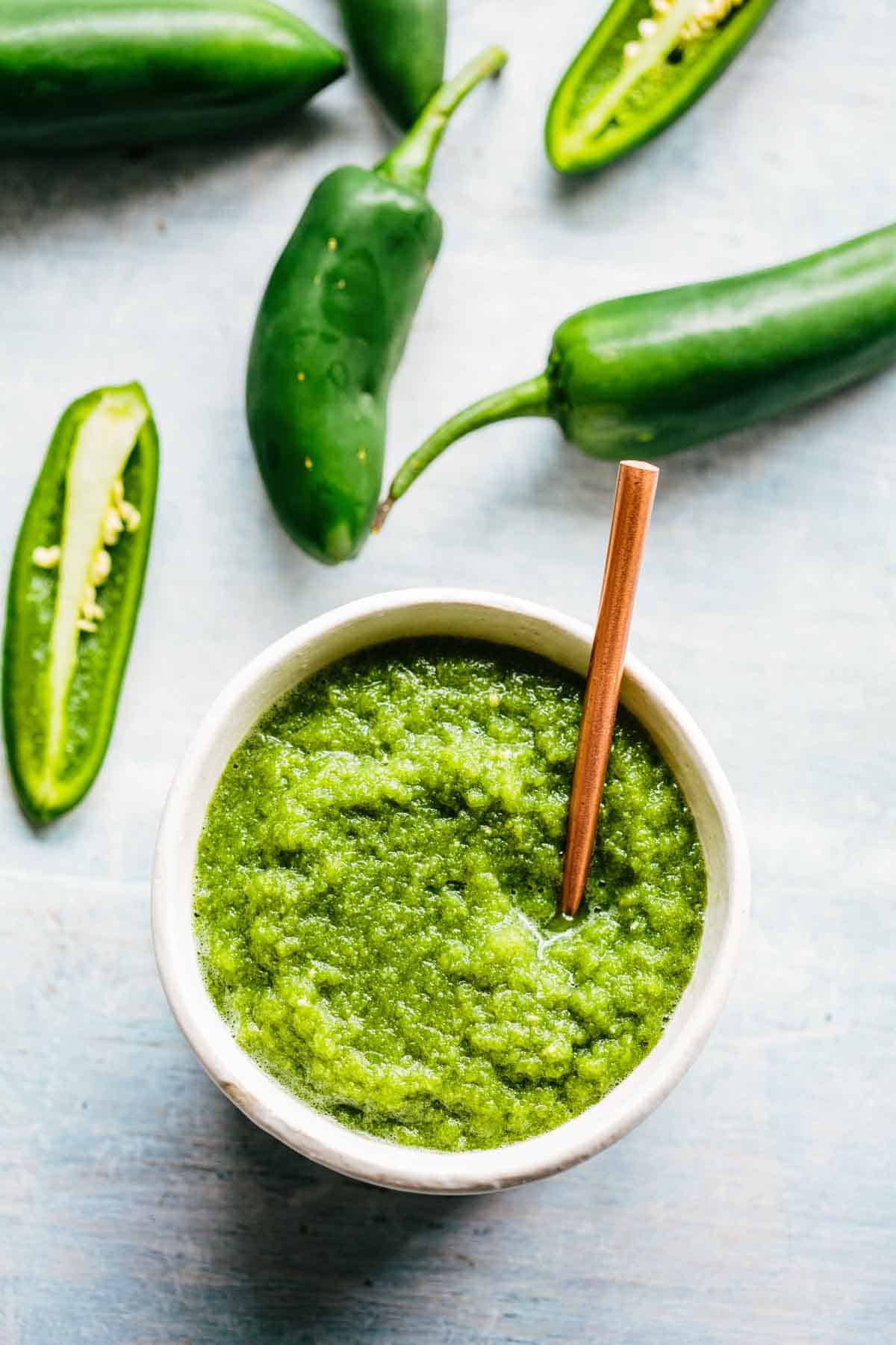 A small bowl of Jalapeño hot sauce with a spoon and jalapeños in the background.