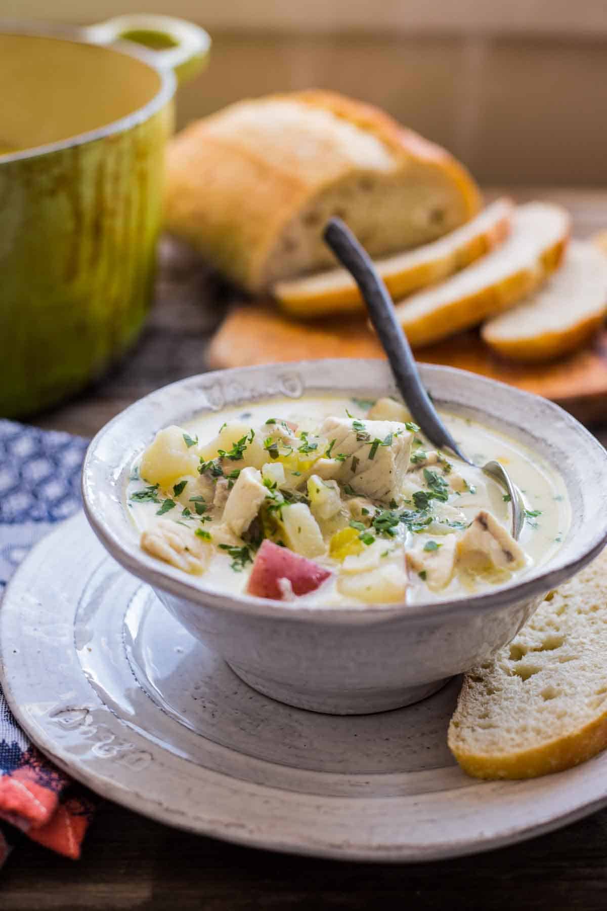 Side view of a bowl of fish soup with a green Dutch oven and loaf of bread in the background.