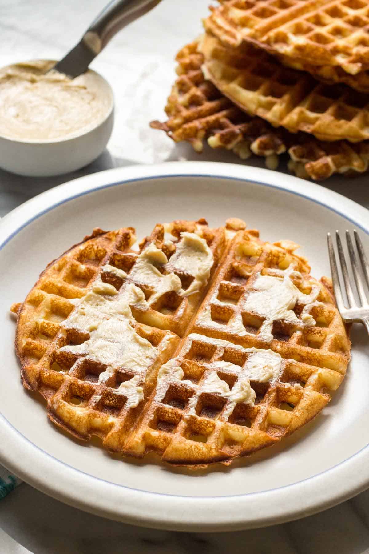 A plate with a waffle with butter smeared on it and a dish of butter and stack of waffles in the background. 