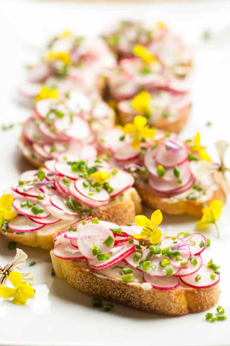 A white platter of toasts topped with thinly sliced radishes, herbs and edible flowers.