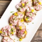Overhead shot of a white platter of buttered radish toasts topped with fresh herbs and edible flowers on top.
