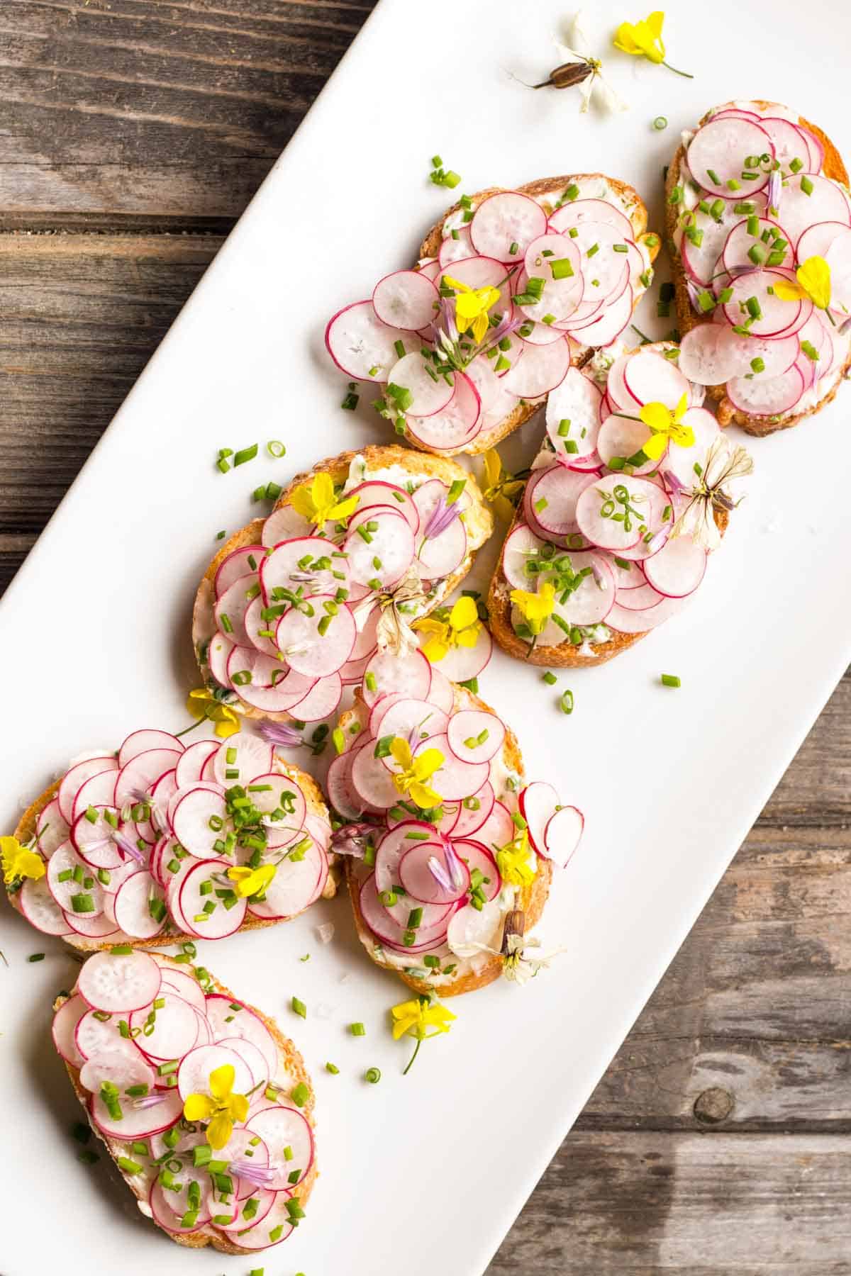 Overhead shot of a white platter of buttered radish toasts topped with fresh herbs and edible flowers on top.