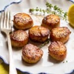 A white plate with pan seared scallops with fresh thyme and a lemon in the background.
