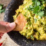 Fingers holding a tortilla chip dipping into a bowl of guacamole.