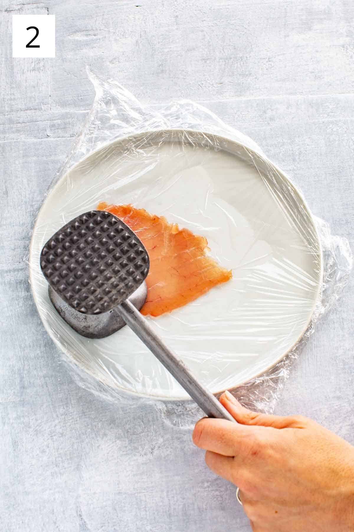 A meat mallet pounding a slice of raw tuna under plastic wrap on a white plate.