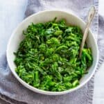 A white bowl of sautéed rapini on a gray cloth.