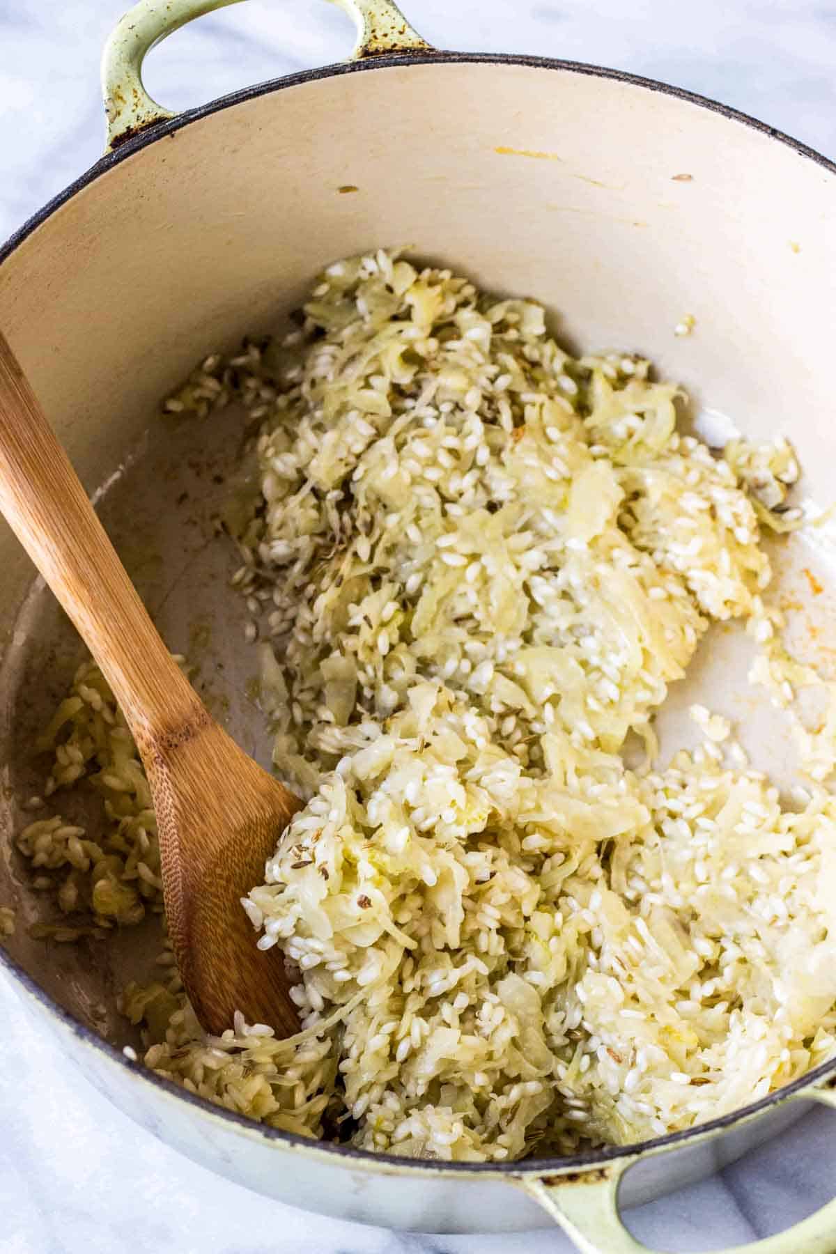 Rice and fennel in a cast iron dutch oven being stirred with a wooden spoon.