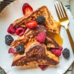 Three triangles of French toast on a plate with maple syrup and berries.