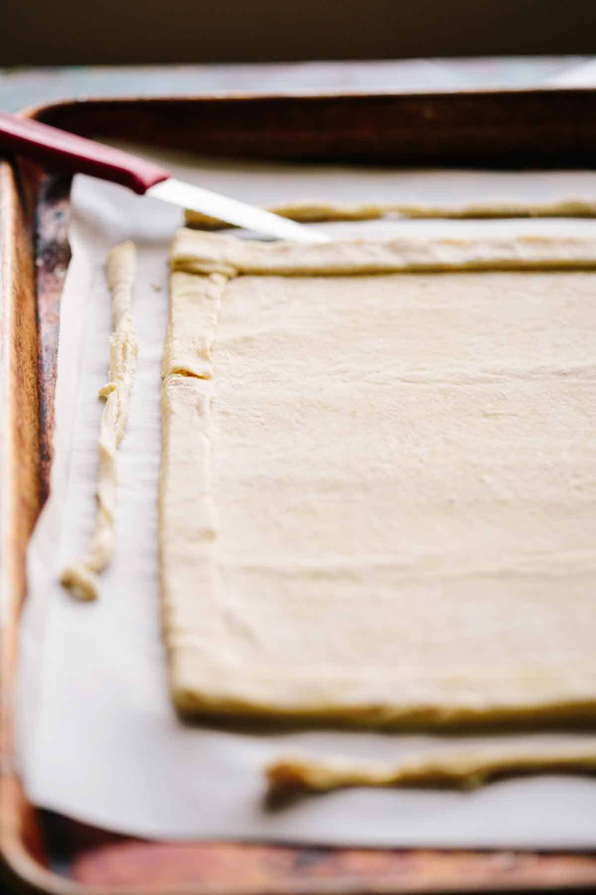 Uncooked puff pastry on a sheet pan with a paring knife cutting the edges.
