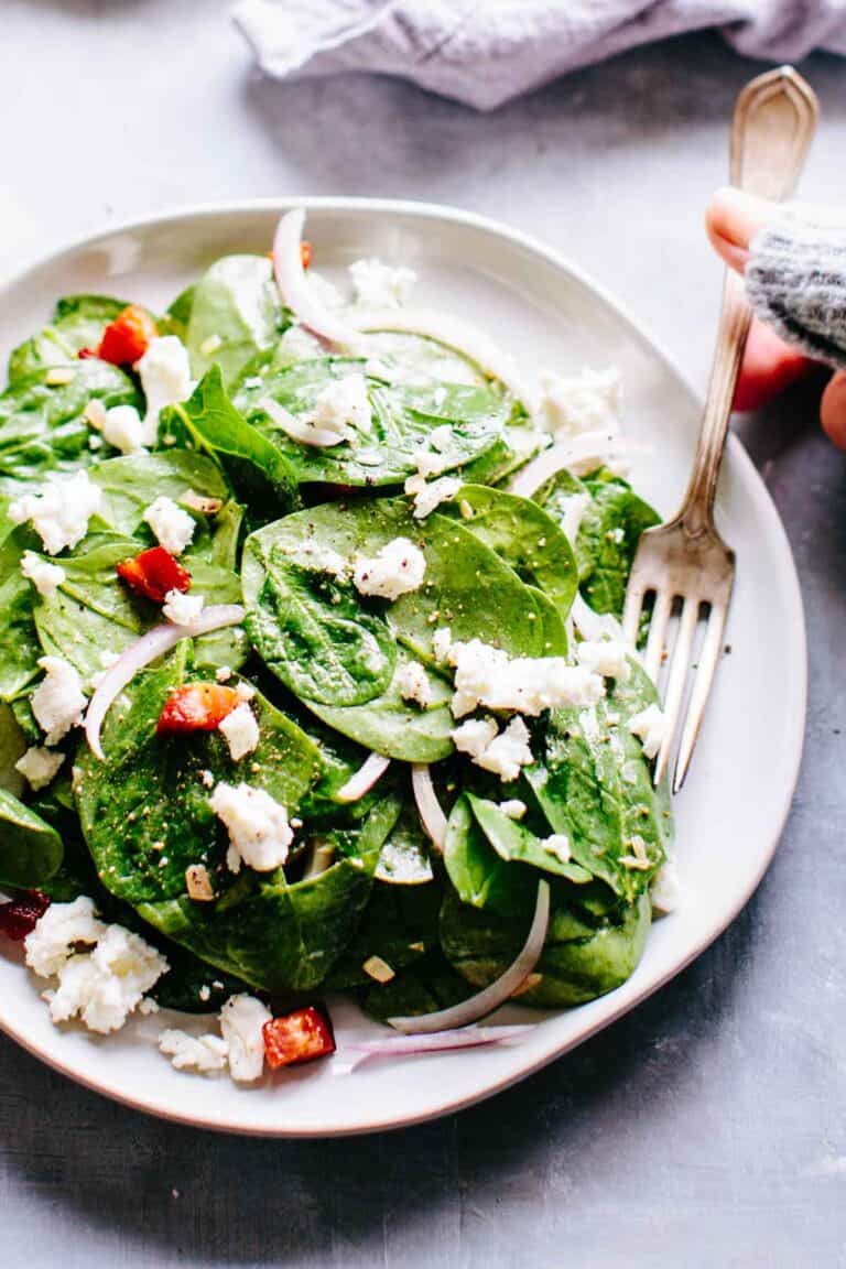 A hand scooping up some spinach leaves with goat cheese, pancetta and red onions with a fork.