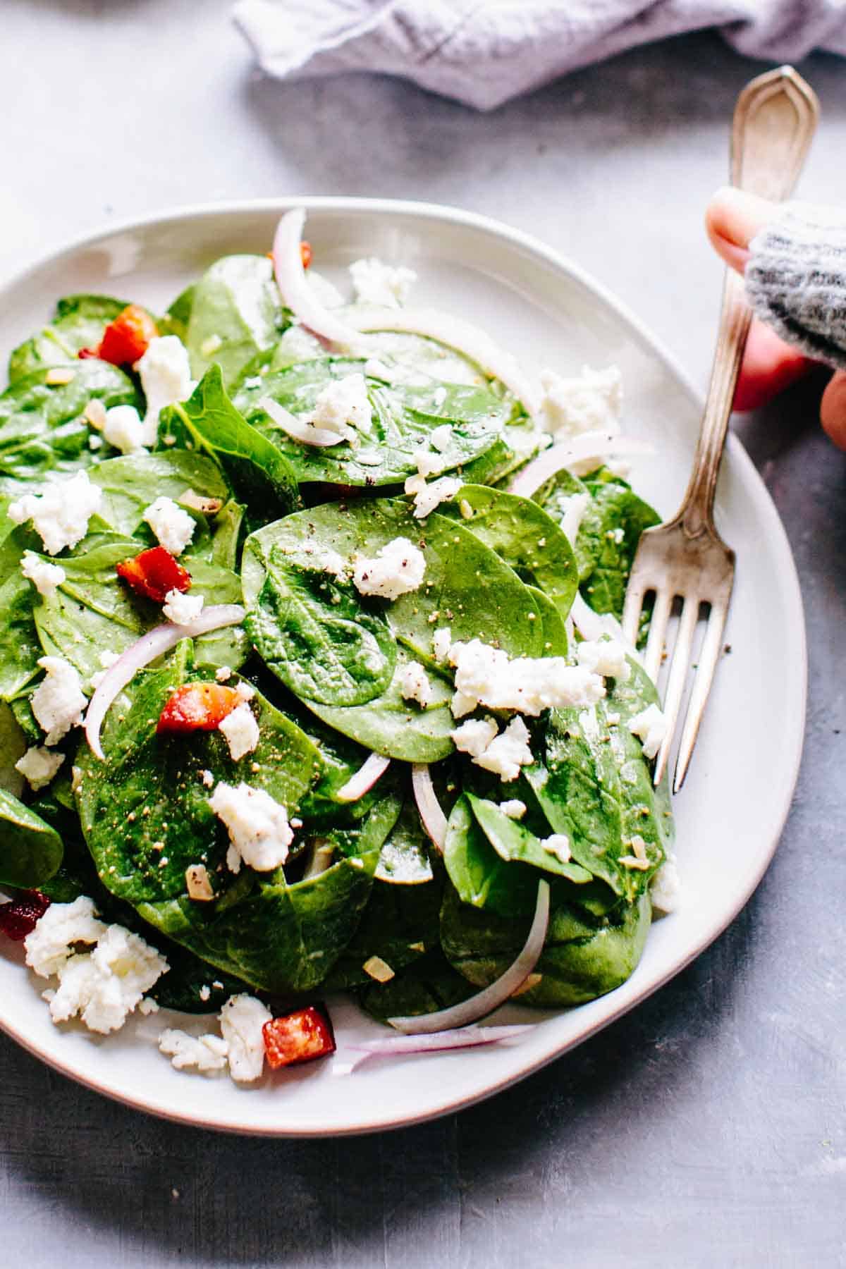 A hand scooping up some spinach leaves with goat cheese, pancetta and red onions with a fork.