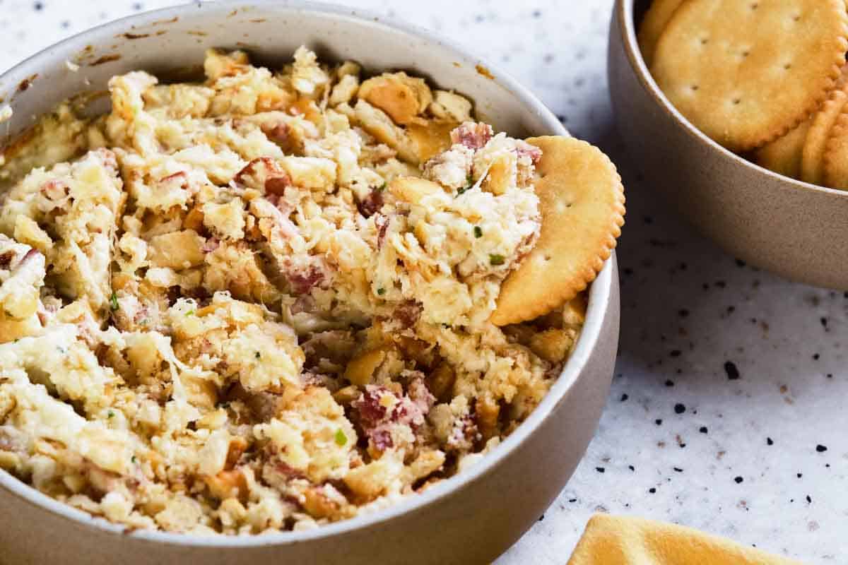 A cracker dipping into a bowl of dip with bacon.
