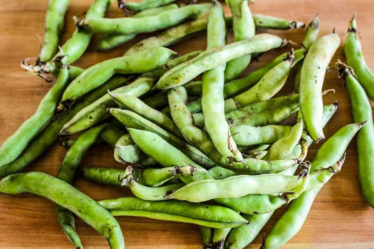Whole fresh Fava beans in their pods on a wooden cutting board.