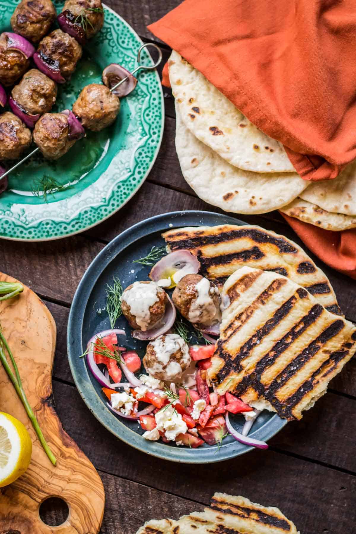 Table spread with lamb meatballs on skewers, a stack of a pita, and a dinner plate including tomato salad.
