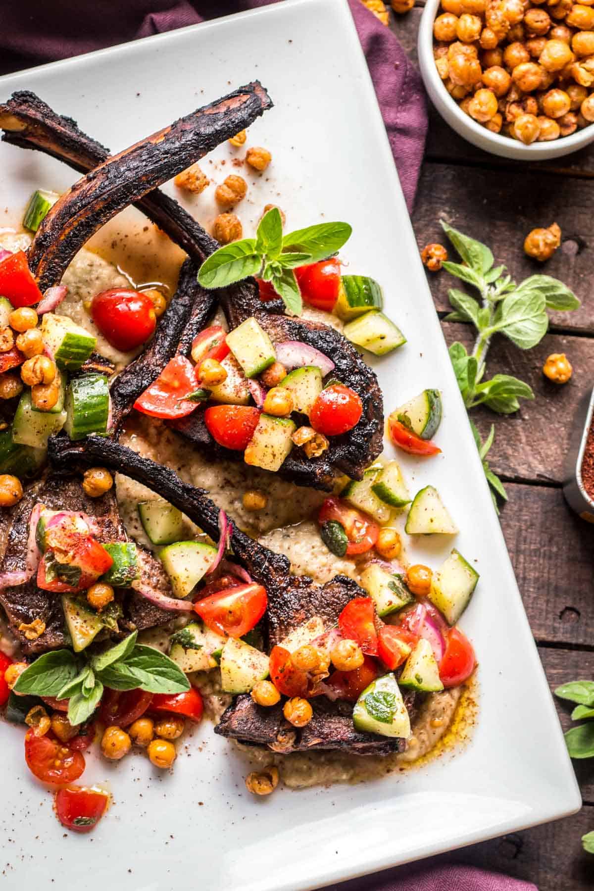 White serving platter with lamb chops covered in chickpea, tomato, cucumber salad.