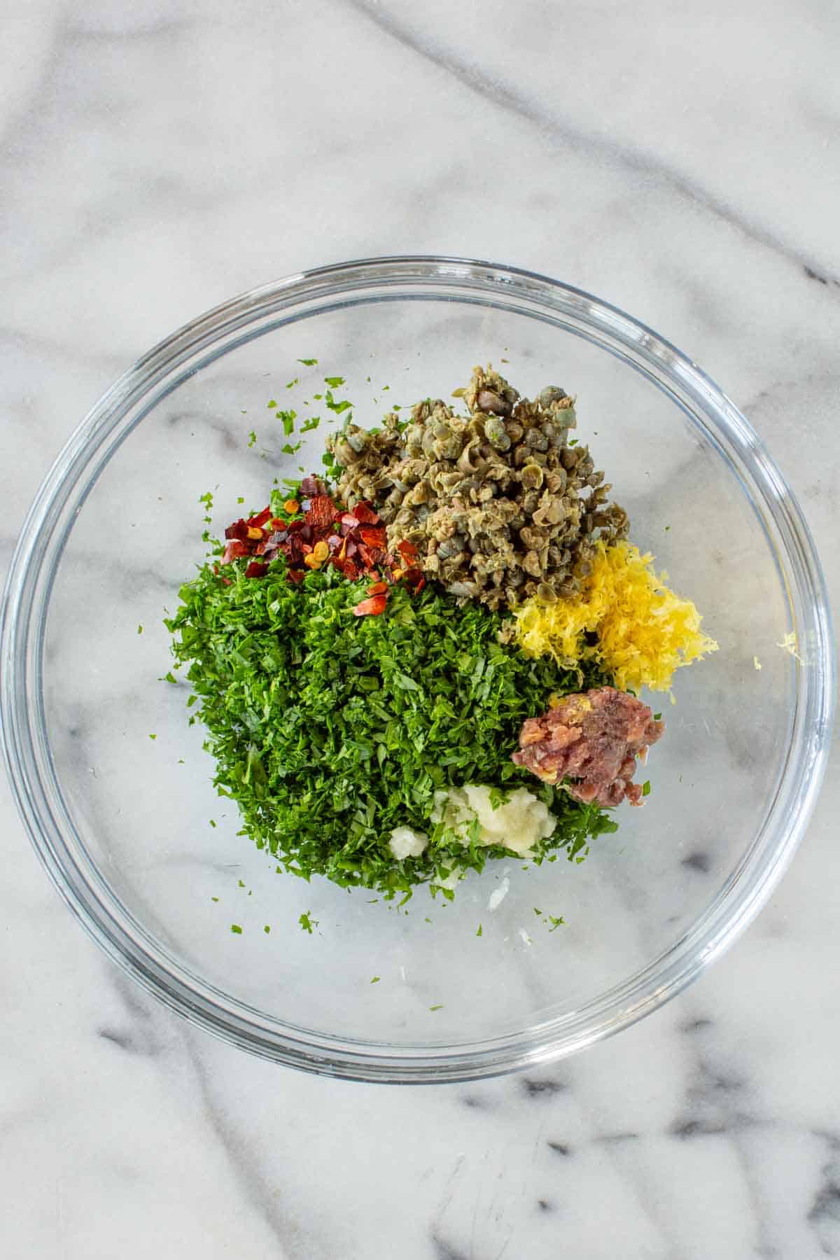 A glass bowl with parsley, anchovies, lemon zest, capers, garlic and chili flakes.